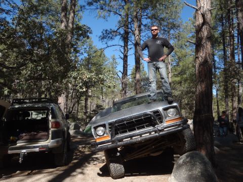 1978 Ford Bronco - A man stands atop a 1978 Ford Bronco as it navigates rocky terrain in a wooded area.