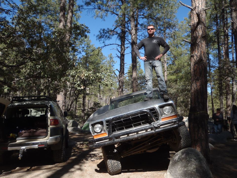 1978 Ford Bronco, silver, tilted on rocks, man standing on roof, forest setting