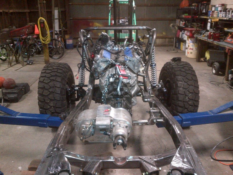 Custom off-road vehicle frame with engine and transmission installed, sitting on lift arms in a garage