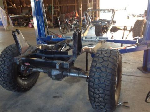 1985 Jeep CJ7 - The frame of a Jeep CJ is suspended on a car lift, showing its chassis and large off-road tires.