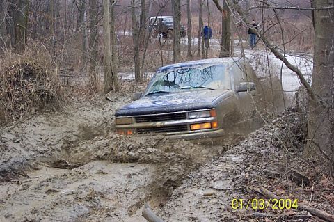 1978 Ford Bronco - An SUV is stuck deep in mud on a wooded trail, with another vehicle visible further back.