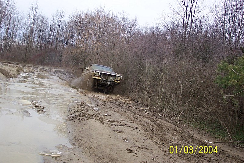Brown 1978 Ford Bronco driving through mud on a dirt road, front 3/4 view