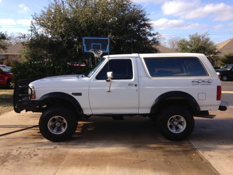 1994 Ford Bronco - This 1994 Ford Bronco is shown from the side, parked on a concrete surface with trees and houses in the background.