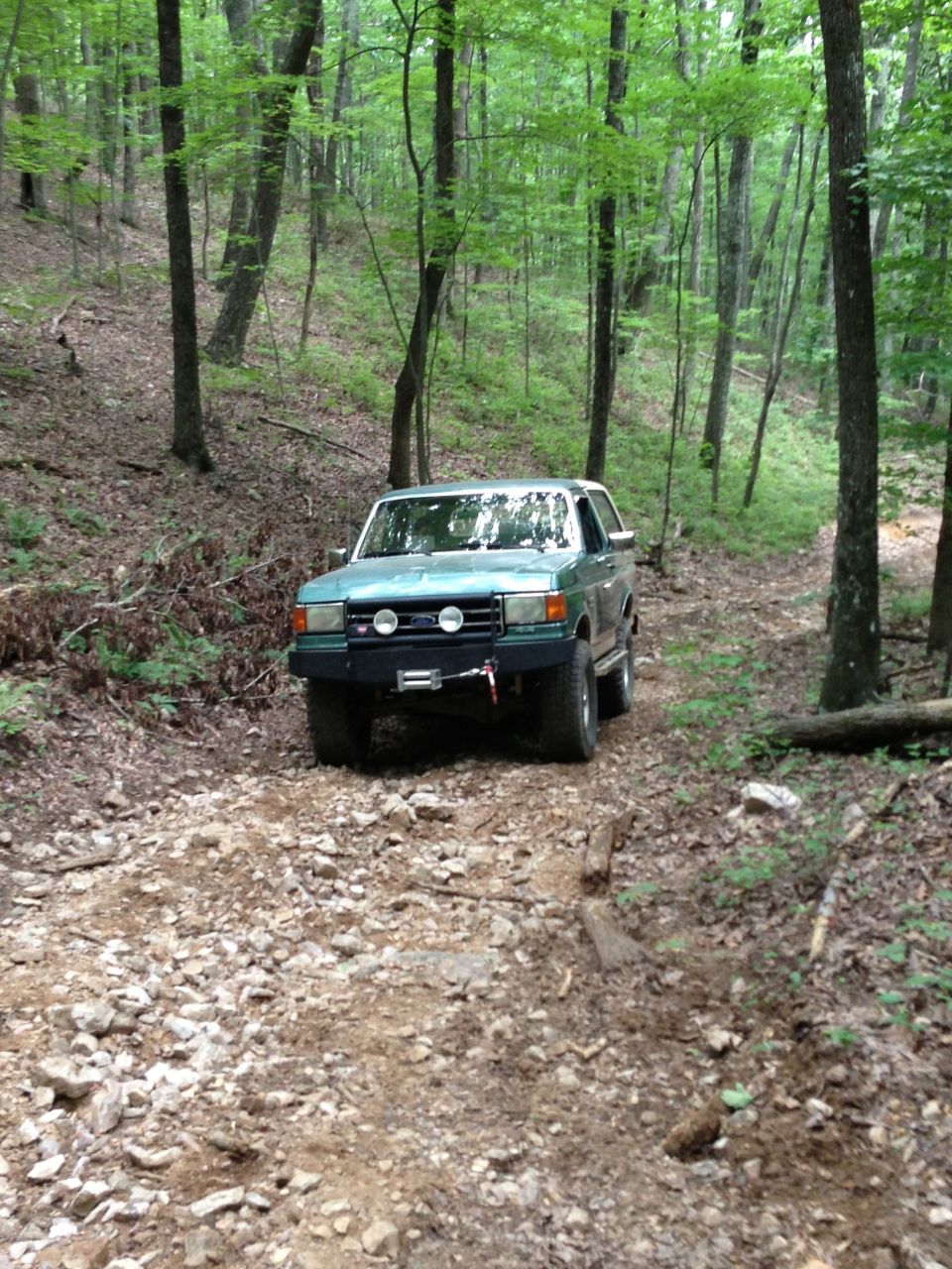 Teal Ford Bronco with aftermarket bumper and winch on a rocky, wooded trail