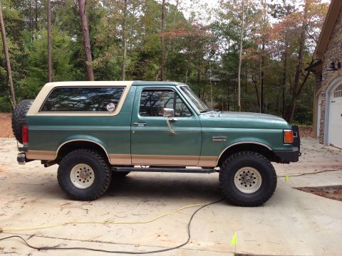 1989 Ford Bronco - Side view of a green and tan Ford Bronco, showing its off-road tires and spare tire carrier.