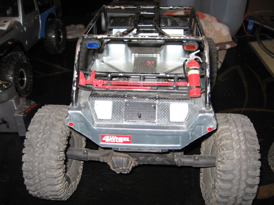 Rear view of a remote control rock crawler with a fire extinguisher and Hi-Lift jack mounted. Another RC vehicle is visible to the left