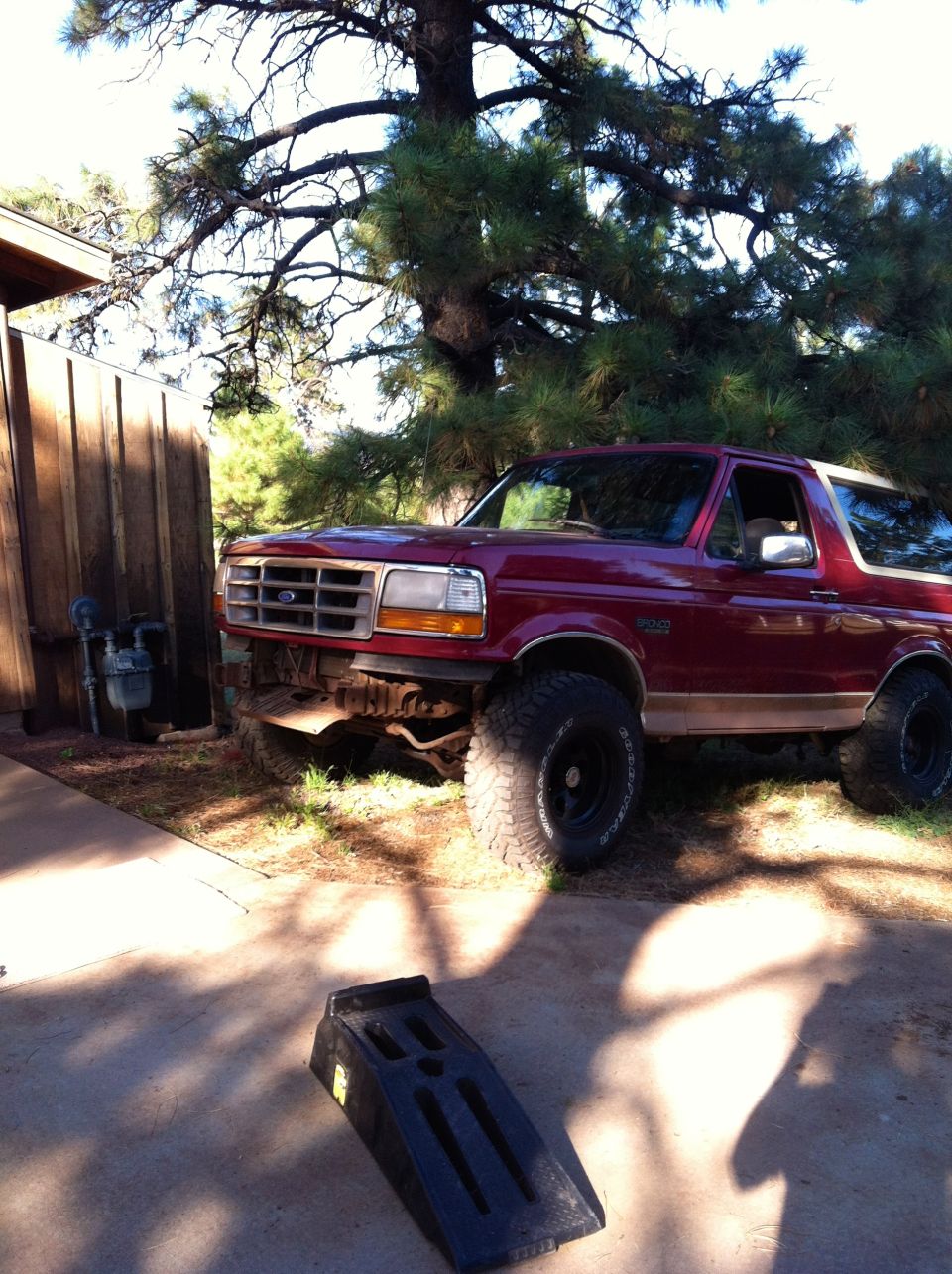 Red Ford Bronco parked next to a wooden fence and a large tree. A car ramp is on the ground in front