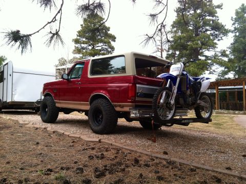 1994 Ford Bronco - A red SUV is parked outdoors with a dirt bike attached to a rear rack and a trailer behind it.