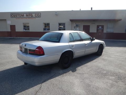 2002 Mercury Grand Marquis - Rear three-quarter view of a silver 2002 Mercury Grand Marquis sedan parked in front of a building with 'CUSTOM MFG. CO.' signage.