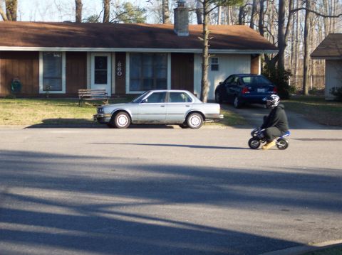 1990 Ford Bronco - A rider in a dark hooded jacket and blue-and-white full-face helmet rides a small blue pocket bike / mini moto down a suburban residential…