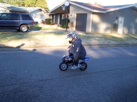 1990 Ford Bronco - A rider in a black hoodie and white full-face helmet rides a small blue pocket bike down a suburban residential street. A dark blue SUV sit…