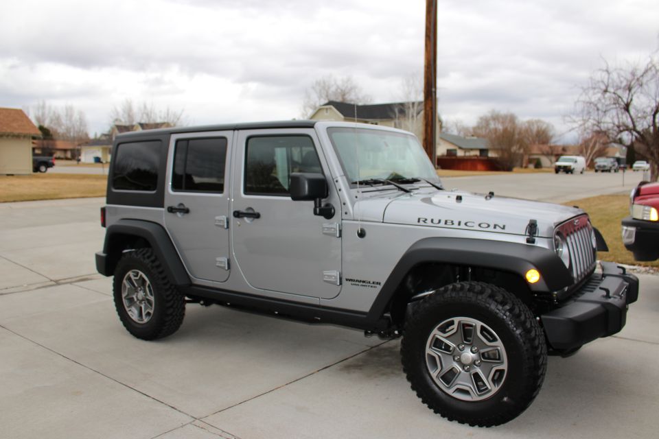 Silver four-door Jeep Wrangler Rubicon with black hardtop parked on a driveway, 3/4 front passenger view