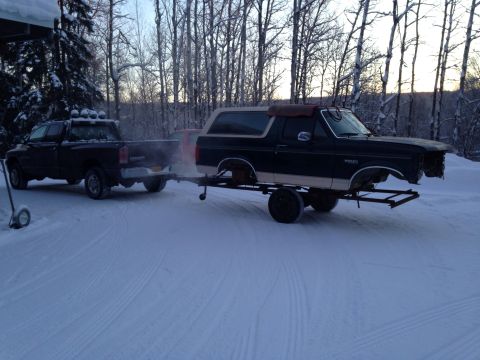 1993 Ford Bronco - Pickup truck tows a 1993 Ford Bronco on a trailer through a snowy landscape.