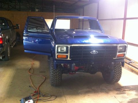 1982 Ford Bronco - A blue Ford Bronco with an aftermarket hood scoop is parked inside a garage, with its driver door ajar.