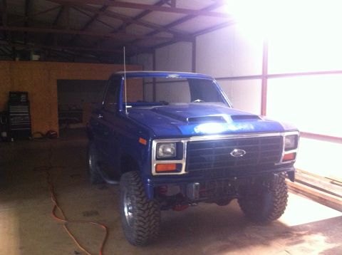 1982 Ford Bronco - Front view of a blue Ford pickup truck with a custom hood scoop and off-road tires inside a garage.