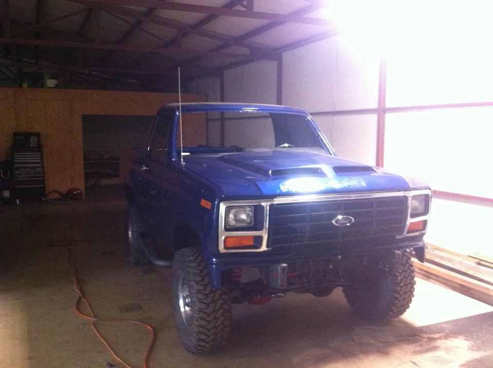Blue Ford pickup truck, likely from the early 1980s, parked indoors. It has a custom hood scoop and large off-road tires