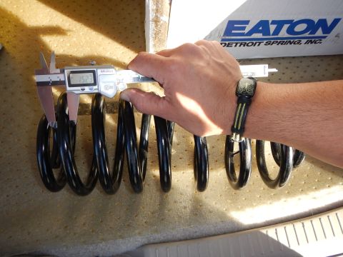 2003 Ford Crown Victoria - hand measures a black coil spring with a digital caliper on a tan pad, with an Eaton Detroit Spring Inc box behind it.
