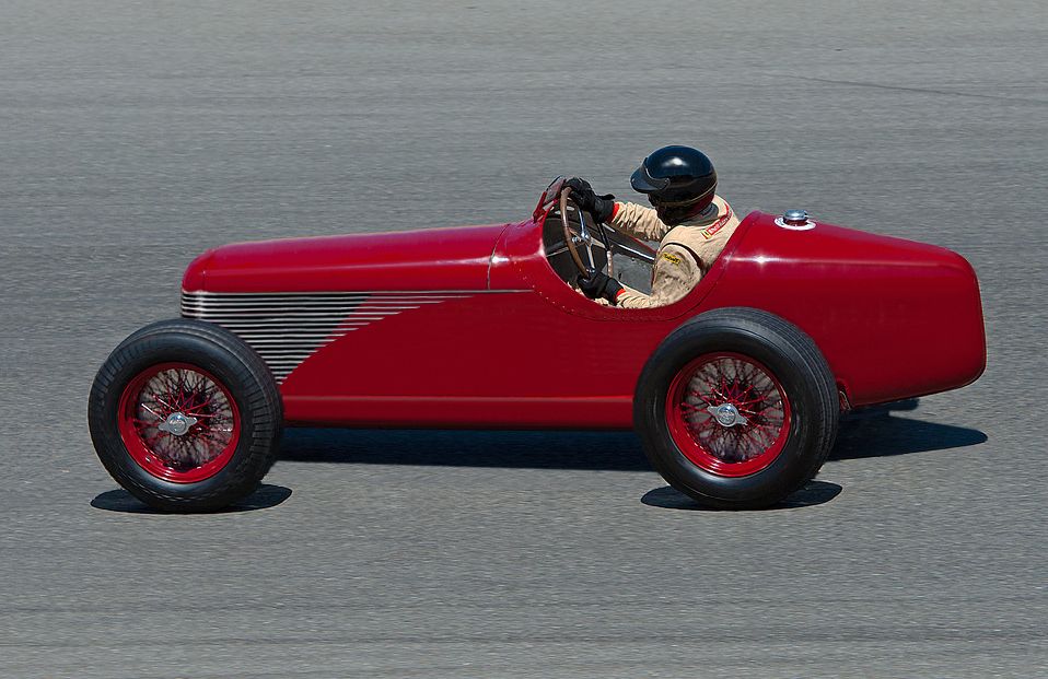 Red vintage open-wheel race car with a driver wearing a helmet and racing suit on a paved track