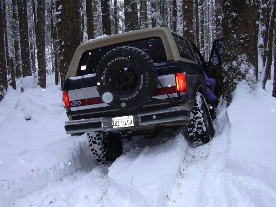 Dark Ford Bronco climbing a snowy forest trail, rear tire lifted, spare on tailgate