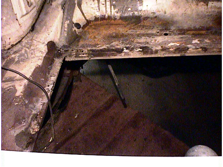 rusted car floor pan with a large hole revealing darkness below, wires visible