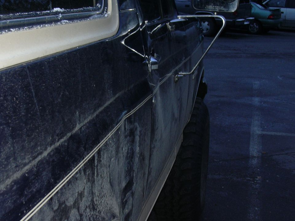 Close-up of a dark truck's door, mirror and rocker panel salted from winter, parking lot behind