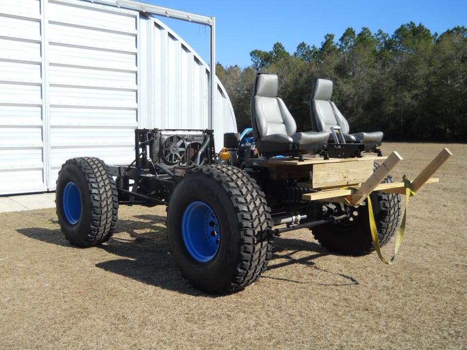 Custom off-road vehicle chassis with two grey seats, large tires with blue rims, and exposed engine components