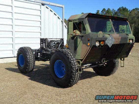 Camouflage military truck chassis with large tires and blue rims, parked outside a building