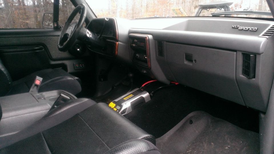 Interior shot of a Ford Bronco, showing the dashboard, steering wheel, seats, and a power inverter on the floor. The 'XLT Bronco' badge is visible on the dash