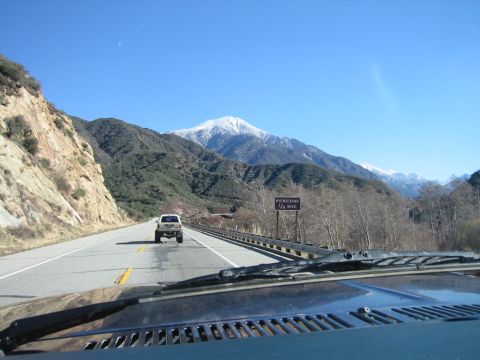 1985 Ford Bronco - A white SUV travels down a highway towards snow-capped mountains under a clear blue sky.