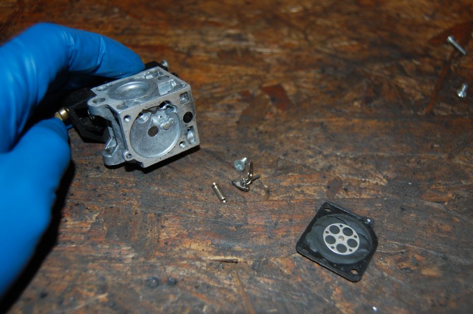Carburetor disassembled on workbench, with blue-gloved hand holding main body