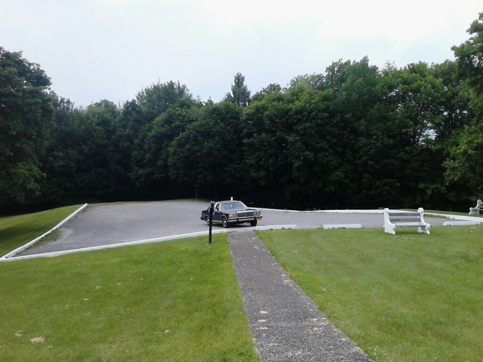 Dark blue Ford Crown Victoria sedan parked alone in a paved lot surrounded by grass and trees