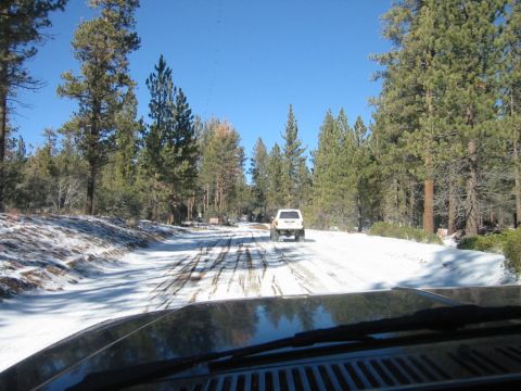 1985 Ford Bronco - Two vehicles travel down a snow-covered road through a forest on a clear day.