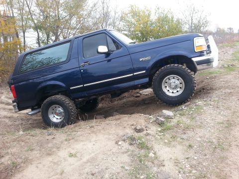 1995 Ford Bronco - A blue 1995 Ford Bronco navigates an off-road trail, demonstrating its capability on uneven terrain.
