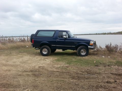 1995 Ford Bronco - A dark blue 1995 Ford Bronco is parked on a grassy bank next to a large body of water under a cloudy sky.