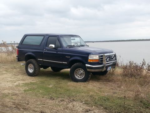 1995 Ford Bronco - A blue Ford Bronco is parked on a grassy area near a large body of water under a cloudy sky.