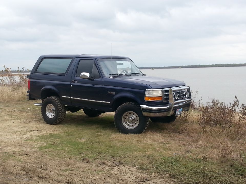 Blue 1995 Ford Bronco parked on grassy shore next to a large body of water