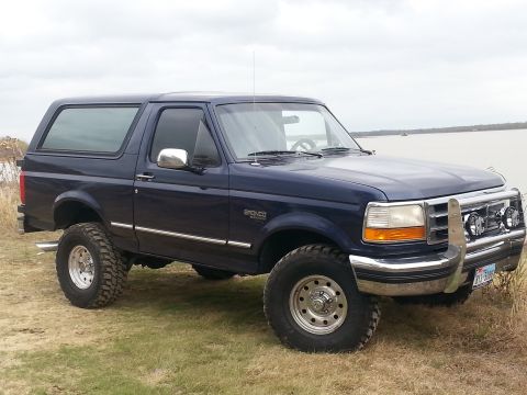 1995 Ford Bronco - A dark blue 1995 Ford Bronco is shown from a three-quarter front view, parked outdoors with a body of water in the background.