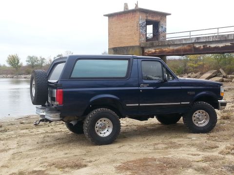1995 Ford Bronco - This 1995 Ford Bronco is seen from a 3/4 front angle, parked near a body of water with a bridge and abandoned structure in the background.