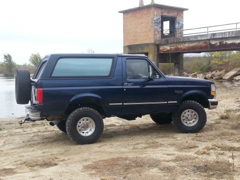 1995 Ford Bronco - 1995 Ford Bronco parked by the water with an abandoned structure in the background.