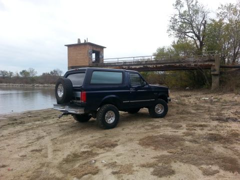 1995 Ford Bronco - A dark blue Ford Bronco is parked on a sandy shore next to a body of water and an old bridge.