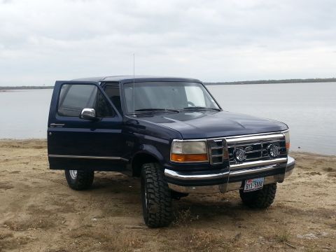 1995 Ford Bronco - This dark blue SUV, likely a 1992 model, is parked on a sandy shore with its driver's door open, facing the water.