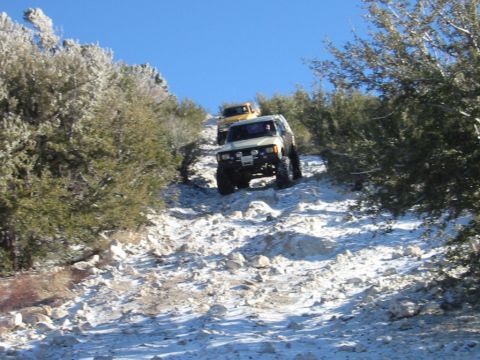 1985 Ford Bronco - Off-road vehicles navigate a challenging, snow-covered mountain path under a clear blue sky.
