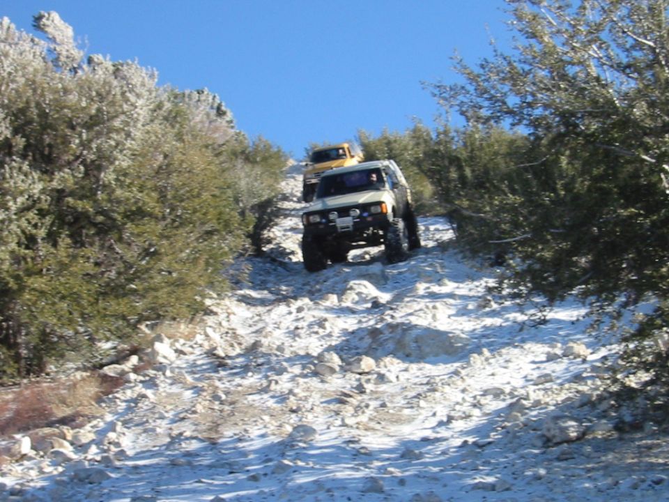 White lifted SUV descending a snowy rocky trail with a yellow vehicle behind it