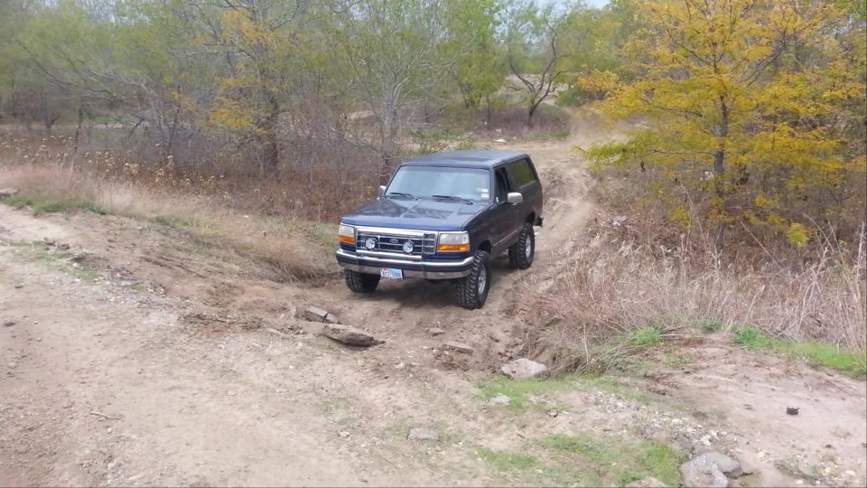 1995 Ford Bronco driving on a dirt trail with trees and dry grass