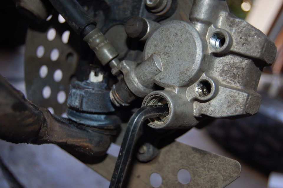 Motorcycle brake caliper with hex wrench inserted, showing perforated disc and suspension components