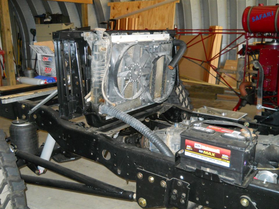 Truck chassis with radiator, fan, and battery installed. Some red machinery is visible in the background