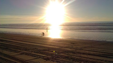 1996 Ford Bronco - A bright sun sets over the ocean as two people walk along a sandy beach with tire tracks.