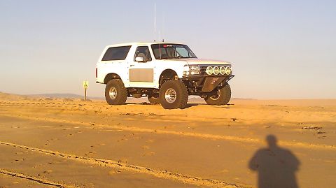 1996 Ford Bronco - A heavily modified white Ford Bronco sits on a sandy terrain under a clear sky.