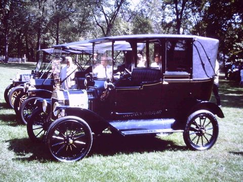 1915 Ford Model T - A line of vintage touring cars, including a dark model with a canvas roof, are parked on a grassy lawn.