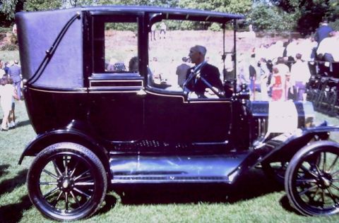 1915 Ford Model T - A 1915 Ford Model T town car, identified by its distinctive body style, is seen from the side with a driver at the wheel.